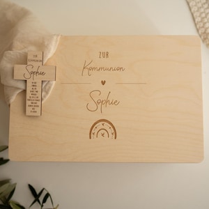 Puede incluir: Una caja de madera de color claro con las palabras "ZUR Kommunion Sophie" y una ilustración de arcoíris. Una cruz de madera con texto está adjunta a la caja. La caja es rectangular con esquinas redondeadas.