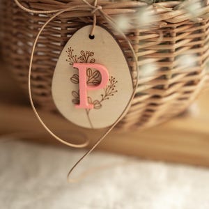 May include: A light brown wicker basket with a wooden egg-shaped tag tied to it with twine. The tag has a floral design and the letter "P" in pink.