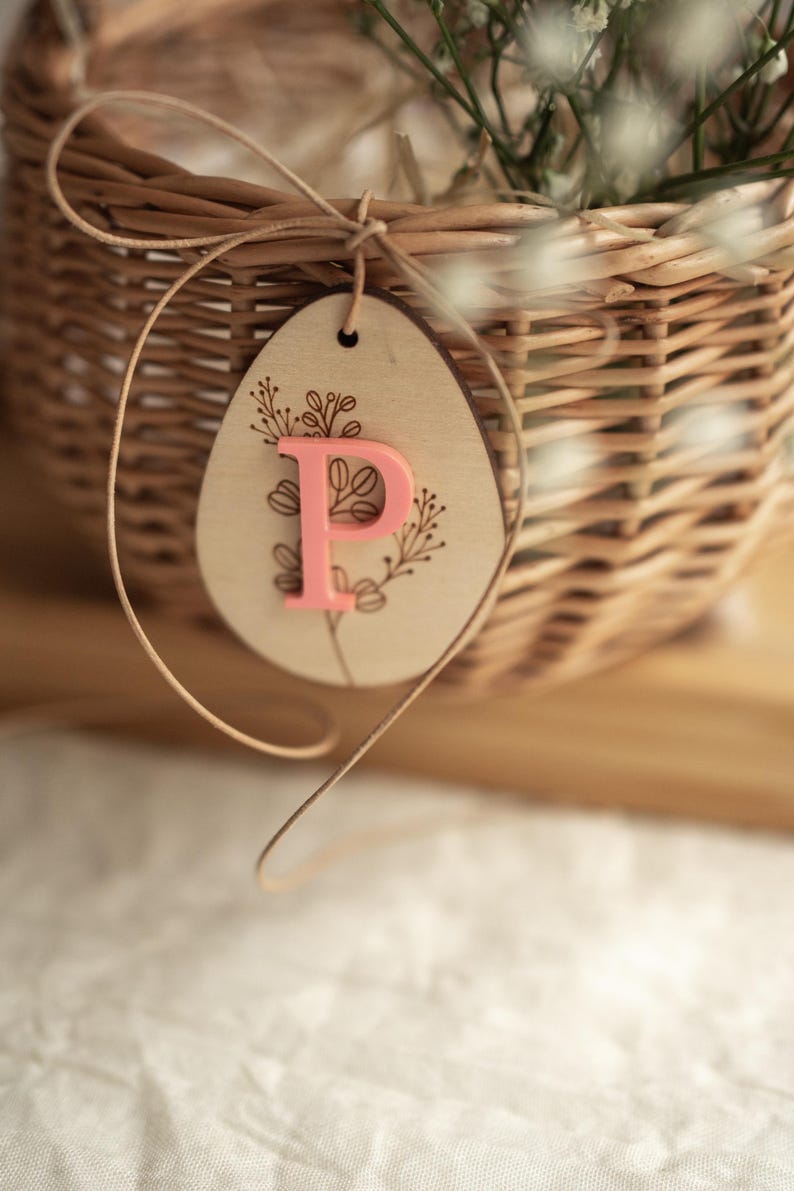 May include: A light brown wicker basket with a wooden egg-shaped tag tied to it with twine. The tag has a floral design and the letter "P" in pink.