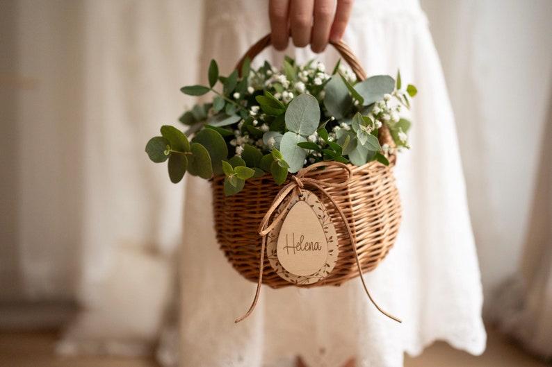 May include: A small wicker basket filled with green eucalyptus and white flowers. A wooden tag with the name "Helena" is attached to the basket.