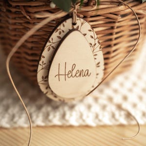 May include: A wooden egg-shaped tag with a floral design and the name "Helena" engraved on it. The tag is tied to a wicker basket with a brown ribbon.