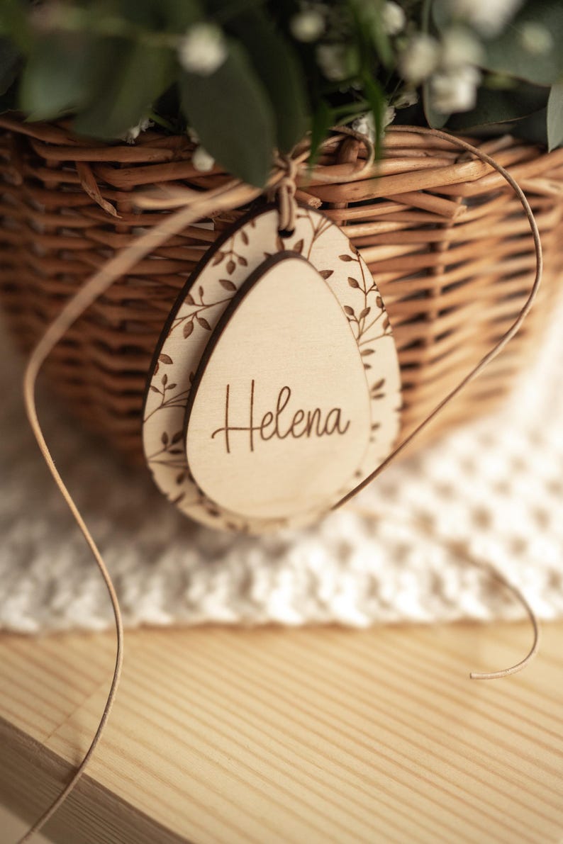 May include: A wooden egg-shaped tag with a floral design and the name "Helena" engraved on it. The tag is tied to a wicker basket with a brown ribbon.