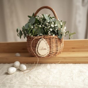 May include: A wicker basket filled with white flowers and greenery, with a wooden egg-shaped tag that says "Helena".