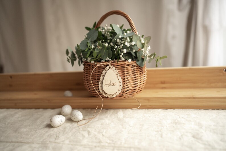 May include: A wicker basket filled with white flowers and greenery, with a wooden egg-shaped tag that says "Helena".