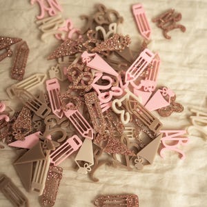 May include: A collection of pink and beige acrylic alphabet letters and pencil shapes with gold glitter accents. The letters are in various sizes and styles.