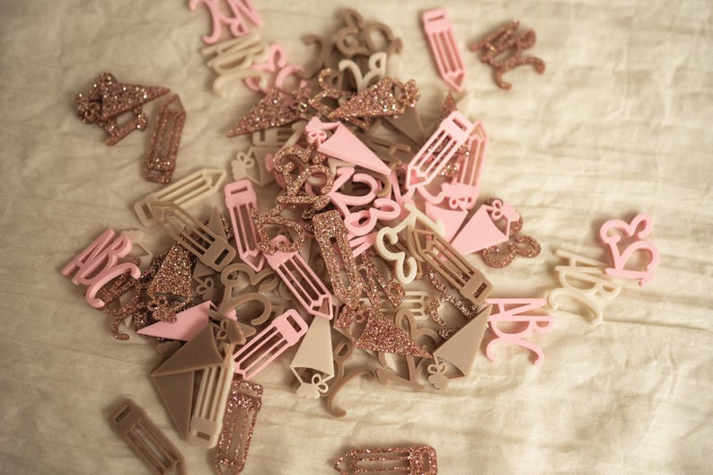 May include: A collection of pink and beige acrylic alphabet letters and pencil shapes with gold glitter accents. The letters are in various sizes and styles.