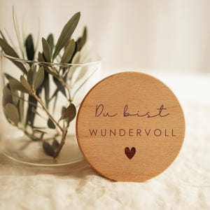 Könnte beinhalten: Ein klarer Glaszylinder mit grünen Olivenzweigen neben einer runden, hellen Holzscheibe. Die Scheibe ist mit dem eingravierten Text "Du bist WUNDERVOLL" in einer Kombination aus Schreib- und Druckschrift sowie einem kleinen Herzsymbol versehen.