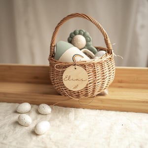 May include: A wicker basket with a wooden tag that says "Elias" is filled with toys. The basket is sitting on a wooden surface with four white speckled eggs in front of it.