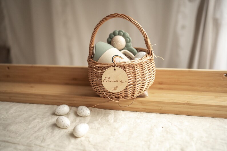 May include: A wicker basket with a wooden tag that says "Elias" is filled with toys. The basket is sitting on a wooden surface with four white speckled eggs in front of it.