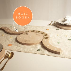 Könnte beinhalten: Ein helles Holz-Tischgesteck mit einer zentralen runden Basis, einer kleinen Glasflasche und einem geschwungenen, segmentierten Bogen. Die Holzoberfläche zeigt kleine weiße Blumeneinlagen und eine Gravur. Eine orangefarbene Grafik mit dem Text HOLZ BÖGEN zeigt auf das Holzbogenelement.