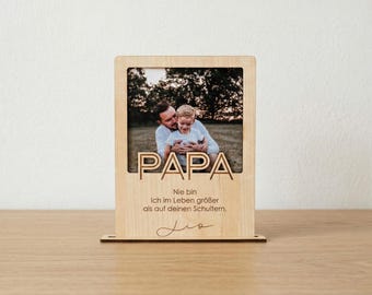 Könnte beinhalten: Ein rechteckiger Bilderrahmen aus hellem Holz mit einem Ausschnitt des Wortes PAPA. Unter dem Foto steht der Text: Nie bin Ich im Leben größer als auf deinen Schultern. Ein handschriftlicher Name, Lio, ist unten eingraviert.