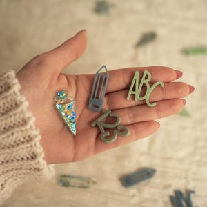 May include: A hand holding various green and blue glitter-covered resin shapes, including a pencil, an ice cream cone, and the letters "ABC".