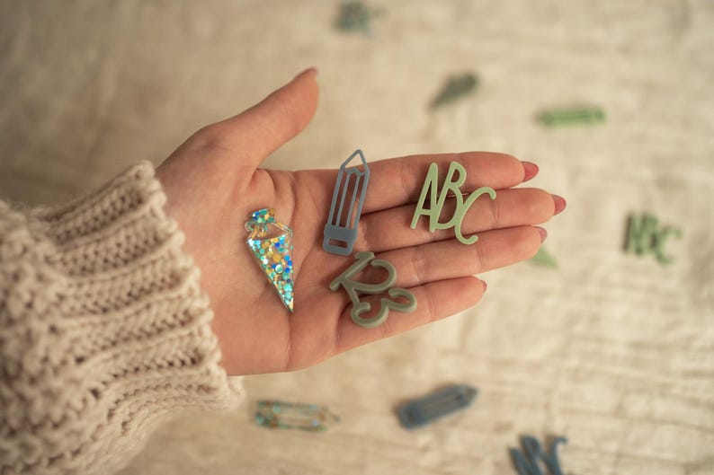 May include: A hand holding various green and blue glitter-covered resin shapes, including a pencil, an ice cream cone, and the letters "ABC".