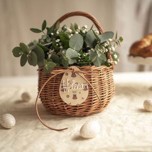 Könnte beinhalten: Ein geflochtener Weidenkorb gefüllt mit Grün und weißen Blumen, mit einem Holzanhänger mit der Aufschrift "Helena". Der Korb steht auf einem Tisch mit gesprenkelten Eiern und einem Brot auf einem weißen Kuchenständer.