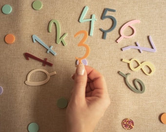 May include: A set of colourful acrylic numbers from zero to nine arranged in a semi-circle, with an orange number three held in the centre. The collection includes various pastel and muted tones, accompanied by small matching circular confetti pieces.