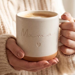 Peut inclure: Une tasse en céramique couleur crème avec une finition mouchetée et une base lisse de couleur terre cuite. Le mot Mama est gravé en écriture cursive sur le devant, accompagné d'un petit cœur gravé sous le texte. La tasse possède une anse.