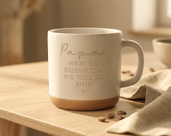 May include: A speckled off-white ceramic mug with a tan base featuring the German text Papa WEIBT DU EIGENTLICH, WIE TOLL DU BIST? and a small heart icon. The mug has a cylindrical body and a curved handle, with a smooth finish and subtle dark flecks.