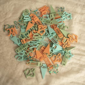 May include: A pile of colorful plastic shapes, including numbers, letters, and pencils, in shades of light blue and orange. The shapes are scattered on a light beige surface.