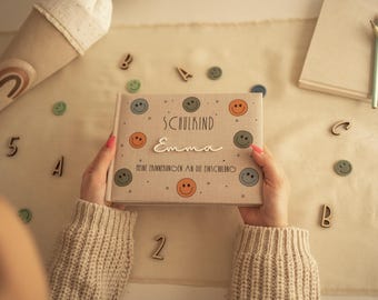 Puede incluir: Un álbum de fotos rectangular de color beige con una cubierta de tela que presenta iconos de caras sonrientes de colores, el texto "SCHULKIND", el nombre "Emma" en letra blanca y el texto "MEINE ERINNERUNGEN AN DIE EINSCHULUNG" en negro.