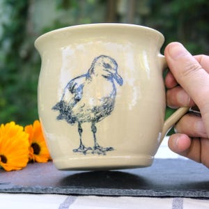 Könnte beinhalten: Eine Keramiktasse mit handgemaltem Design einer Möwe in Blau und Weiß. Die Tasse hat einen abgerundeten Körper und einen Henkel.