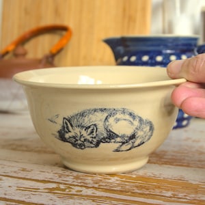 Könnte beinhalten: Eine cremefarbene Keramikschüssel mit einem blauen und weißen Fuchsdesign. Der Fuchs ist zusammengerollt und schläft. Die Schüssel steht auf einer weißen Holzoberfläche.