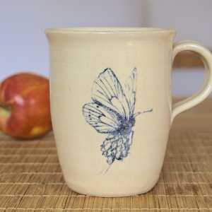 Könnte beinhalten: Eine cremefarbene Keramiktasse mit einem blauen Schmetterlingsdesign. Die Tasse hat einen abgerundeten Griff und einen breiten Boden.