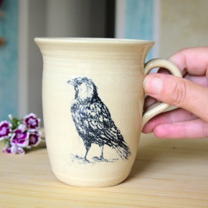Può includere: Una tazza in ceramica color crema con un'illustrazione in bianco e nero di un uccello. La tazza ha un corpo arrotondato e un manico.
