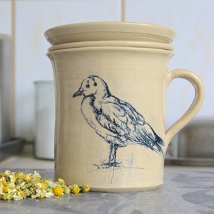 Pode incluir: Uma caneca de cerâmica de cor creme com uma ilustração de um pássaro azul e branco. A caneca tem uma alça e uma tampa.