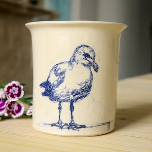 Op de afbeelding: Crèmekleurige keramische keukengerei houder met een blauwe illustratie van een zeemeeuw. De cilindrische houder heeft een licht uitlopende rand en een getextureerd oppervlak. De zeemeeuw is afgebeeld in een gedetailleerde, artistieke stijl.