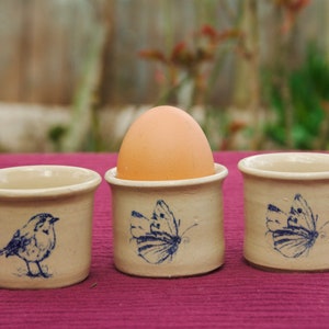 Op de afbeelding: Drie keramische eierdopjes met blauwe en witte ontwerpen. Een dopje bevat een bruin ei. De ontwerpen omvatten een vlinder en een vogel.