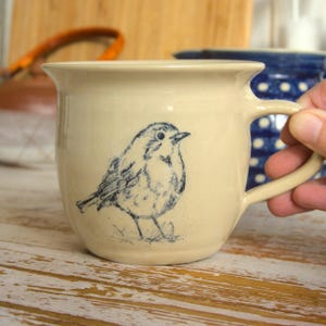 Könnte beinhalten: Eine cremefarbene Keramiktasse mit einer blauen Rotkehlchen-Illustration. Die Tasse hat einen abgerundeten Körper und einen Henkel.