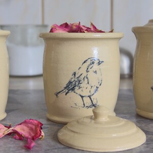 Könnte beinhalten: Cremefarbene Keramikbehälter mit Deckeln, jeweils mit einer detaillierten blauen Vogelillustration verziert. Ein Behälter ist offen und mit rosa Rosenblättern gefüllt. Das Set enthält einen Deckel und zwei weitere Behälter.