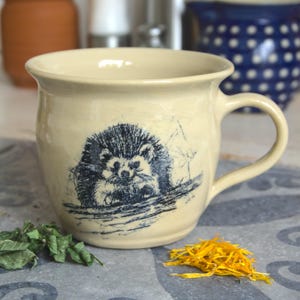 Può includere: Una tazza in ceramica beige con un'illustrazione blu e bianca di un riccio su un ramo. La tazza ha un corpo arrotondato e un manico.