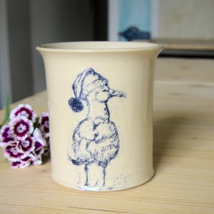 May include: A cream-coloured ceramic mug with a blue illustration of a duck wearing a hat. The duck is facing right and has a fluffy body.