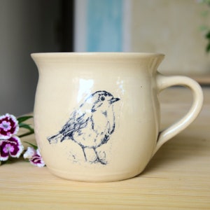 Könnte beinhalten: Eine cremefarbene Keramiktasse mit einer Blauvogel-Illustration. Die Tasse hat einen abgerundeten Körper und einen Henkel.