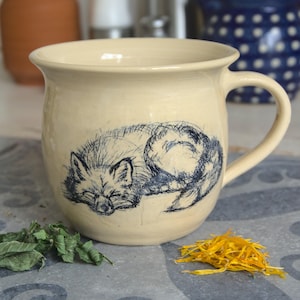 Può includere: Una tazza in ceramica color crema con un'illustrazione blu e bianca di una volpe addormentata. La tazza ha un corpo arrotondato e un manico.