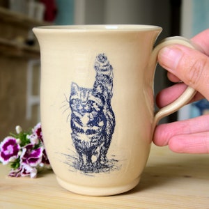 Könnte beinhalten: Eine beige Keramiktasse mit einer blauen Illustration einer Katze, die ihren Schwanz hochhält. Die Tasse hat einen abgerundeten Griff.