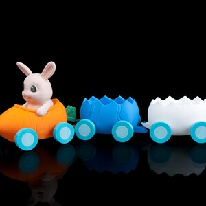May include: A whimsical Easter-themed toy train set. The train features a carrot-shaped engine with a bunny, followed by blue and white eggshell cars, all with light blue wheels. The set is designed with a glossy finish against a black background.