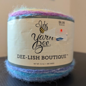 May include: A colorful Yarn Bee Dee-Lish Boutique yarn skein. The label features a bee logo and the text "DEE-LISH BOUTIQUE". The yarn is a blend of blue, purple, and yellow hues. Net weight is 5.3 oz, with 590 yards of yarn.