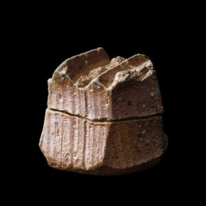 Może przedstawiać: Małe, ręcznie robione, brązowe ceramiczne pudełko z pokrywką. Pudełko ma teksturowaną powierzchnię z pionowymi liniami i szorstką, nierówną górną krawędzią. Pudełko jest ustawione na czarnym tle.