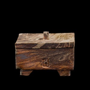 Puede incluir: Una pequeña caja de madera cuadrada con tapa con bisagras y cuatro patas cortas. La caja está hecha de madera con un patrón de vetas arremolinadas en tonos marrones y beige. Un pequeño asa rectangular está en la parte superior de la tapa.