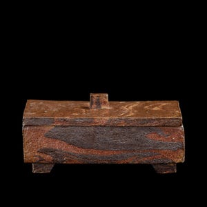 Op de afbeelding: Een rechthoekige, handgemaakte keramische doos met een deksel en kleine voetjes. De doos heeft een getextureerd, gemarmerd uiterlijk met tinten bruin en rood. Bovenop het deksel zit een kleine, vierkante knop.