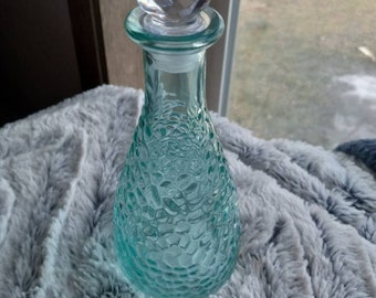 May include: A teal blue glass bottle with a textured surface and a clear crystal stopper. The bottle is sitting on a gray fuzzy surface.