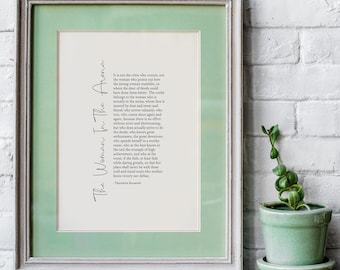 Op de afbeelding: Een ingelijst afdruk met een groene passe-partout en een witte achtergrond. De afdruk bevat een citaat van Theodore Roosevelt over de vrouw in de arena. Het citaat is in zwarte tekst en staat gecentreerd op de pagina. De lijst is lichtgrijs.