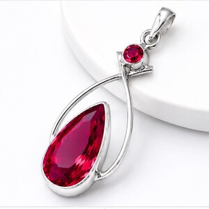 May include: A silver-toned pendant featuring a large, pear-shaped, deep red faceted gemstone set within a curved metal frame. Above the main stone, a smaller, round, deep red faceted gemstone is set in a triangular metal mount with a bail.