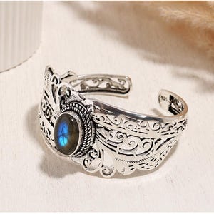 Pode incluir: Uma pulseira de punho prateada com uma pedra preciosa oval azul iridescente no centro. A pulseira apresenta intrincados desenhos vazados e é marcada com "925". A pedra preciosa é rodeada por uma borda de contas.