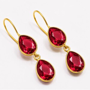 May include: A pair of gold-toned drop earrings featuring two faceted, pear-shaped deep red crystals each. The earrings are suspended from curved gold-toned ear wires, with the crystals set in smooth gold-toned metal frames that catch the light.