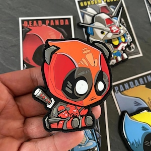 May include: A colorful patch featuring a Deadpool-inspired panda design, holding a katana. The patch is predominantly red and black, with white eyes and gray accents. Other patches with cartoon characters are visible in the background.