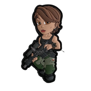 Op de afbeelding: Een cartoon patch met een vrouwelijke soldaat die een geweer met vizier vasthoudt. Het personage heeft bruin haar, een zwarte tanktop en een groene en zwarte camouflagebroek. De patch heeft een zwarte rand.