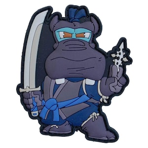 May include: A cartoon patch of a ninja hippo in purple, blue, and grey. The hippo is holding a katana and a spiked weapon. The patch has a black border and is set against a white background.